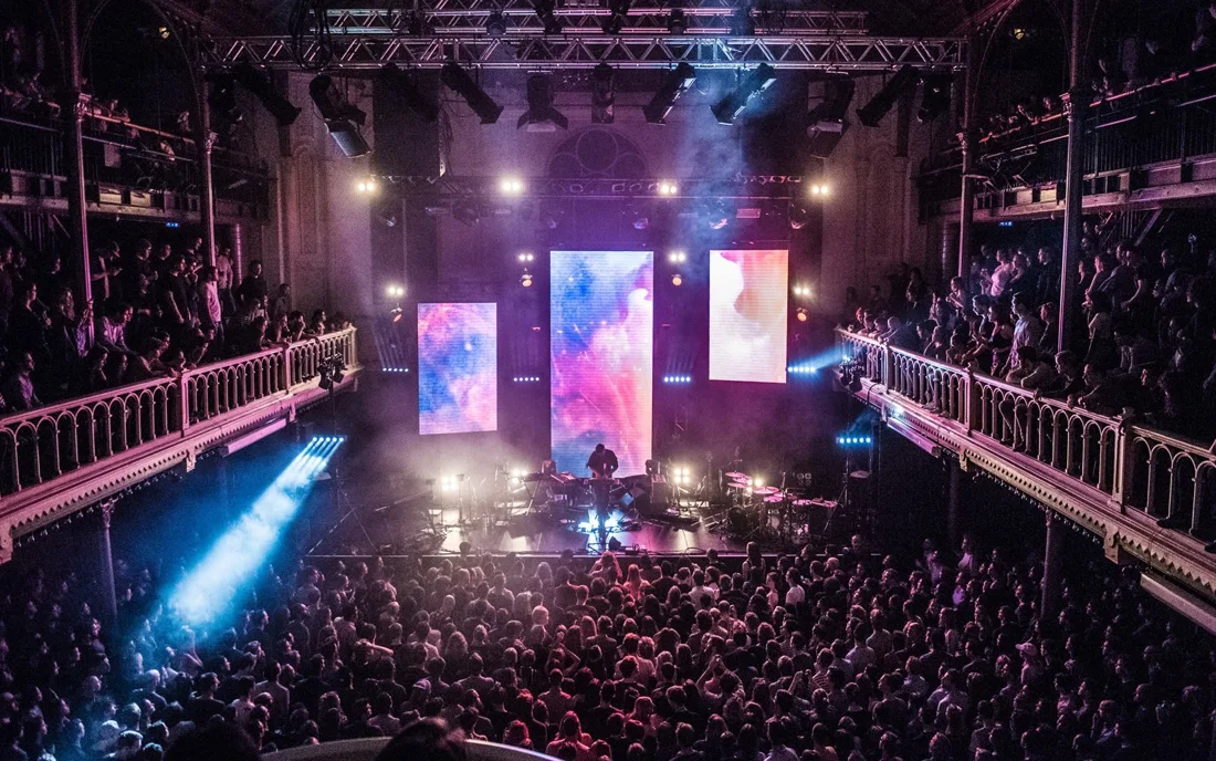 A concert crowd and performers beneath stage lights inside Paradiso, with both balconies visible